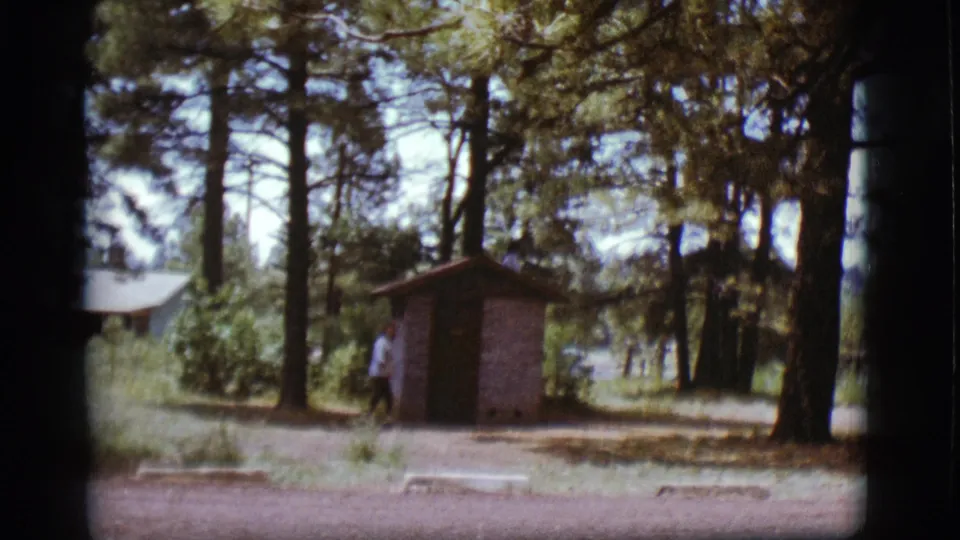 1968 home-movie frame showing a woman walking away from a shed in a pine-covered Flagstaff setting.