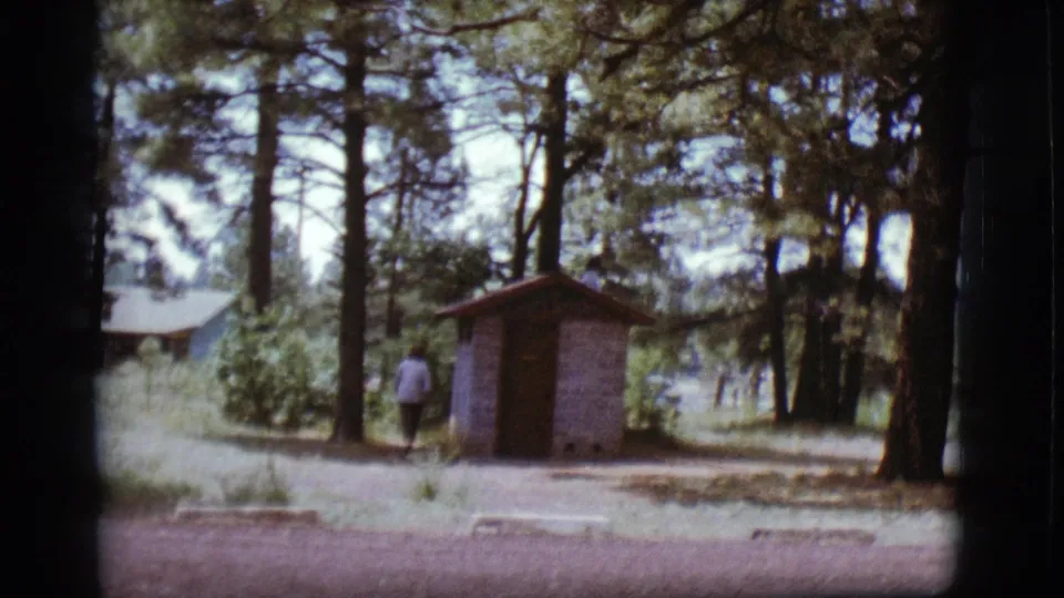 1968 Flagstaff home-movie frame showing a person near a small brick outhouse in a pine campground clearing.