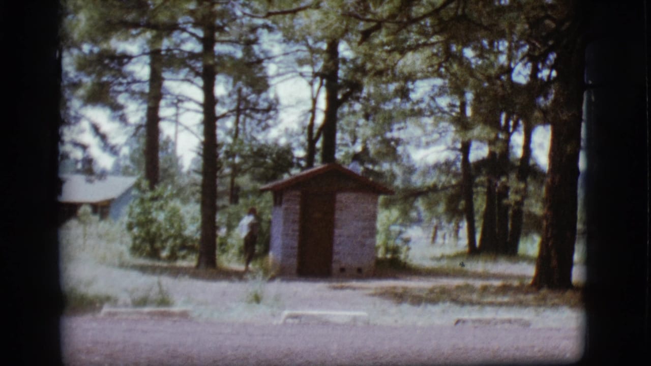 Flagstaff, Arizona campground view with a small brick outhouse, pine trees, dry grass, and a distant person.