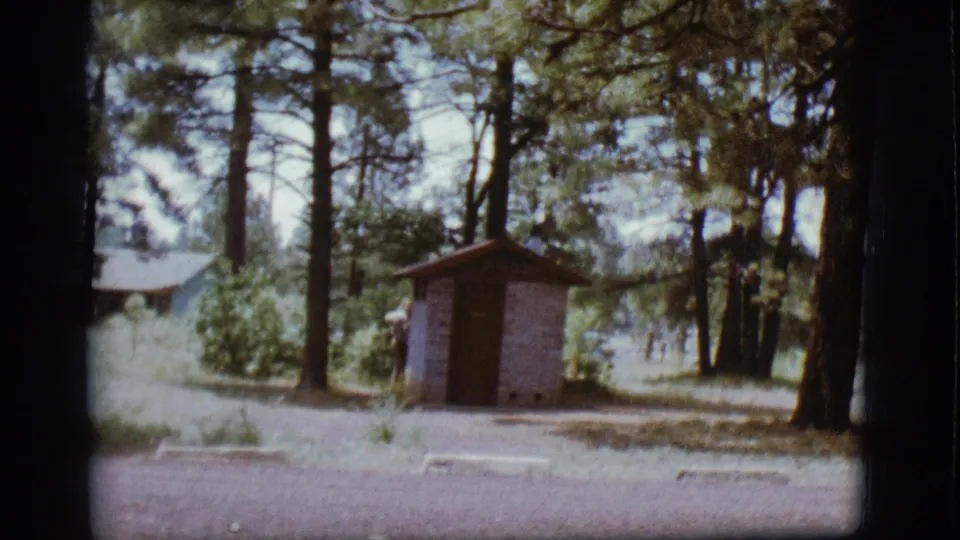 1968 8mm frame of a person partly obscured by a small outhouse in a Flagstaff pine campground.