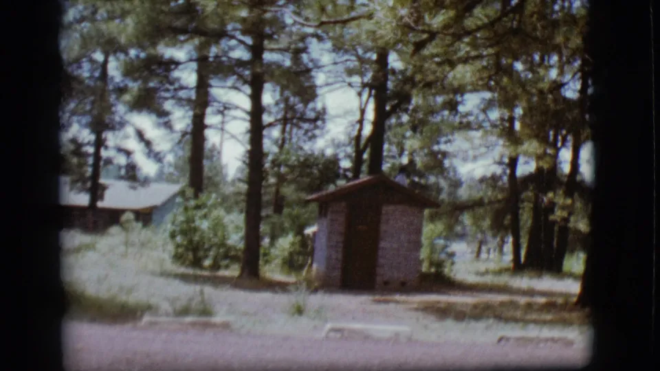 Flagstaff campground frame with a small brick outhouse centered among pines and a person partly visible behind it.