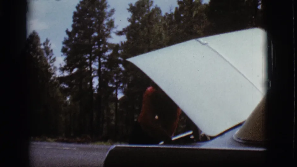 The open hood of a light-colored car fills the frame, with a man partly obscured behind it in the Flagstaff campsite footage.