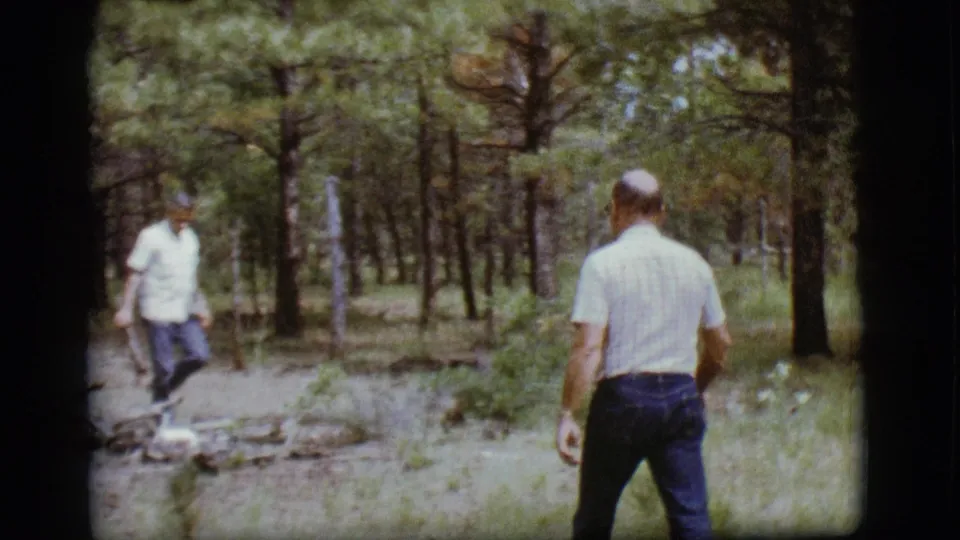 Two adult men walk through a pine forest area in the 1968 Flagstaff home-movie clip, one seen from behind in a striped shirt.