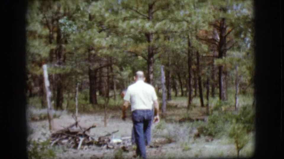 1968 home-movie frame near Flagstaff showing a man walking toward a small branch pile in a sunlit pine clearing.