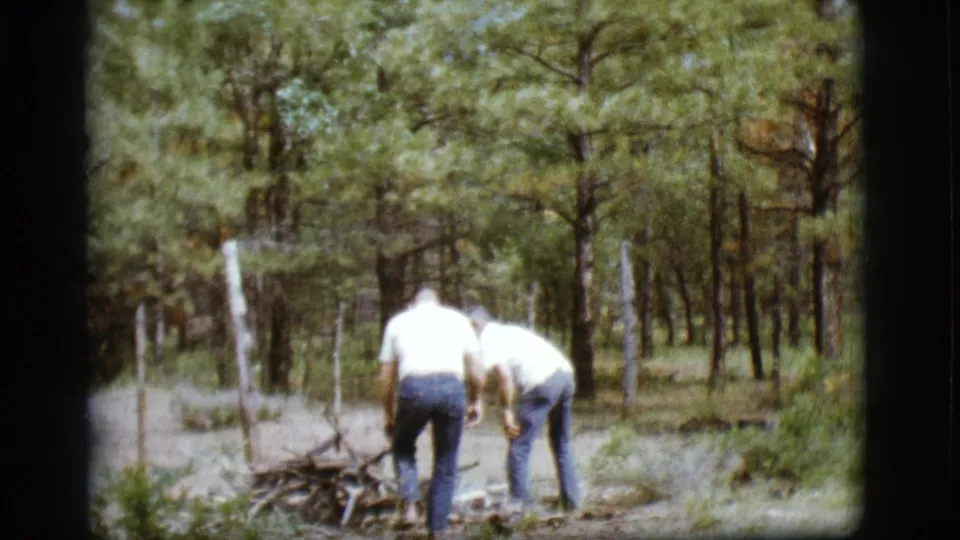 1968 Flagstaff-area frame showing two older men bending near firewood in a pine forest clearing on 8mm film.