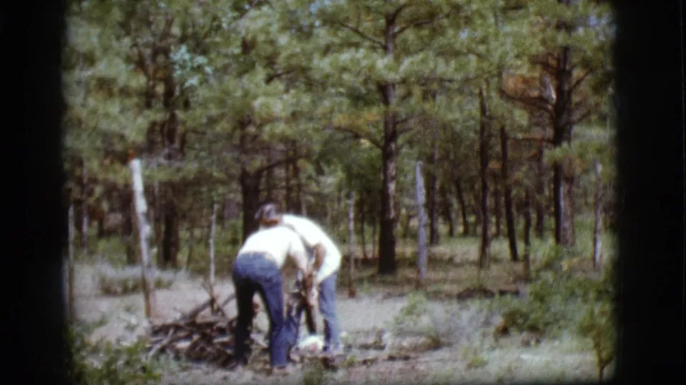 Two adult men bend over a small pile of dry sticks in a wooded Flagstaff setting, with fence posts behind them.