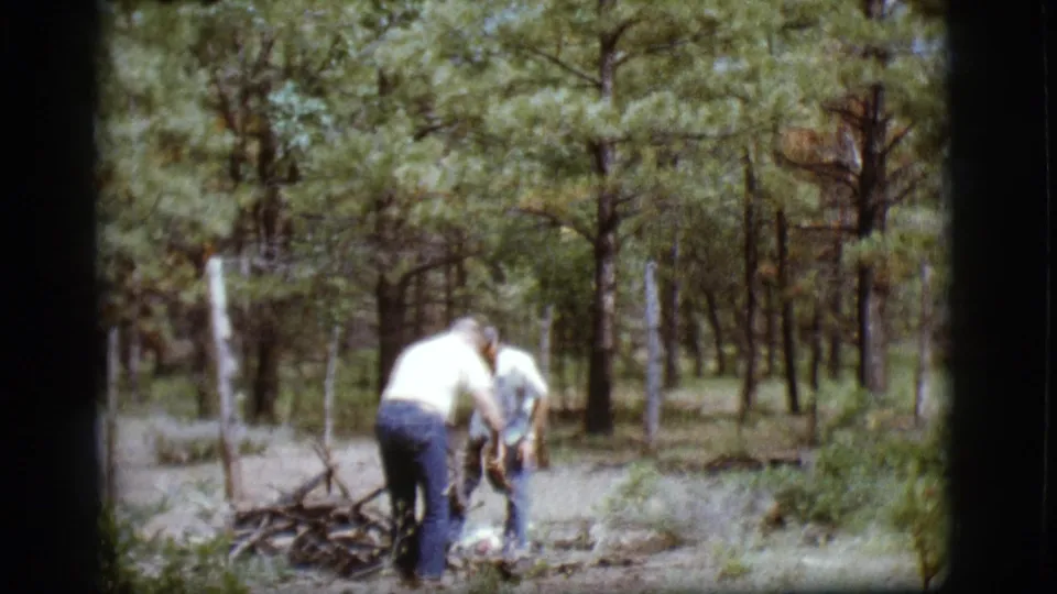 In 1968 Flagstaff, Arizona home-movie footage, two adults gather sticks among pine trees near a branch pile.