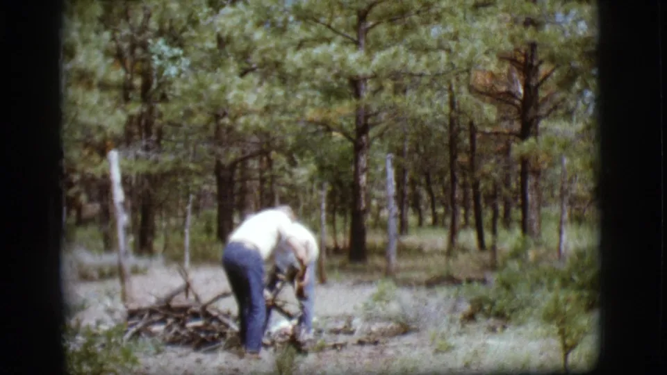 An adult in a light shirt bends over branches in a pine forest setting near Flagstaff, Arizona, in 1968 home-movie footage.