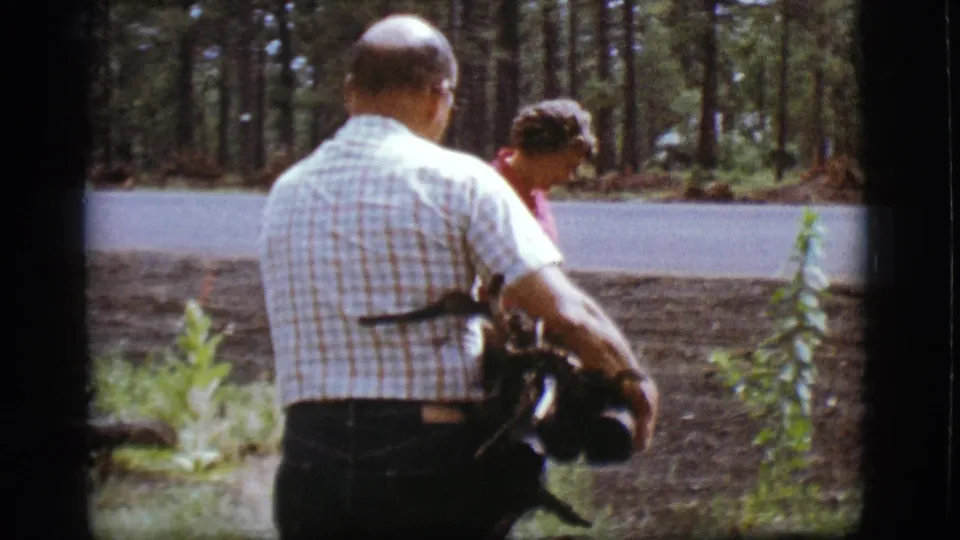 A man in a plaid shirt joins the woman, holding firewood near the forest road in the 1968 Flagstaff, Arizona footage.