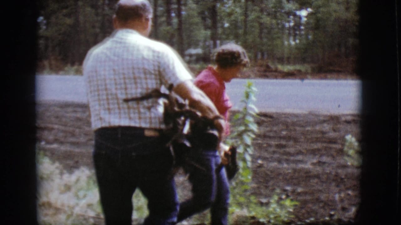 Two adults carry sticks near a forest road, seen from behind on 8mm film.