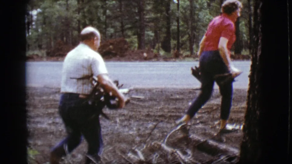 The woman crosses a rough patch of roadside forest ground as the man trails behind with kindling in the 1968 home movie.