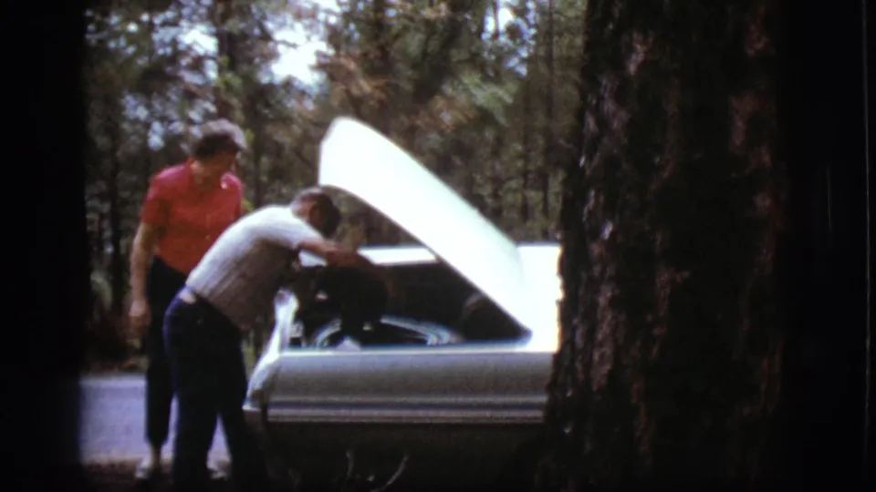 In 1968 Flagstaff footage, a man leans into an open car engine while a woman stands nearby on a forested roadside.