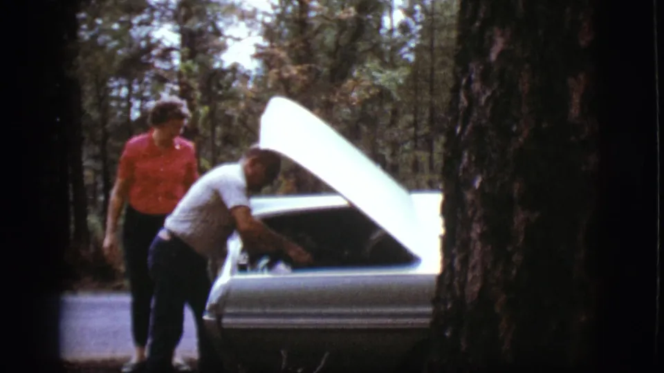 The man continues working under the raised hood of a classic car as the woman watches in the Flagstaff roadside setting.