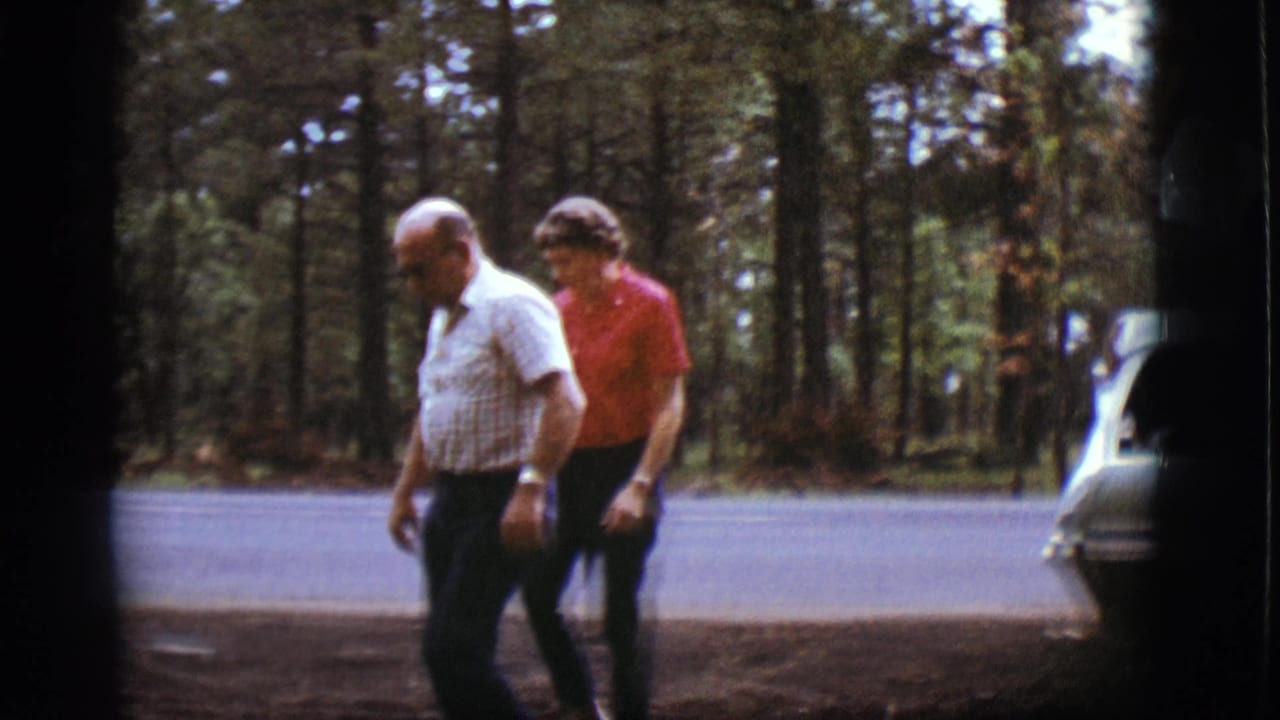Older man and woman walking beside a forest road with pine trees behind them.