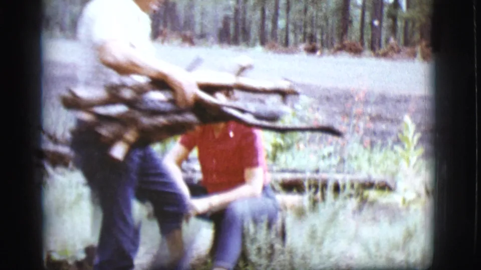 A person carries a bundle of sticks near a forest road while a woman in a red shirt stands close by in the Flagstaff home-movie scene.