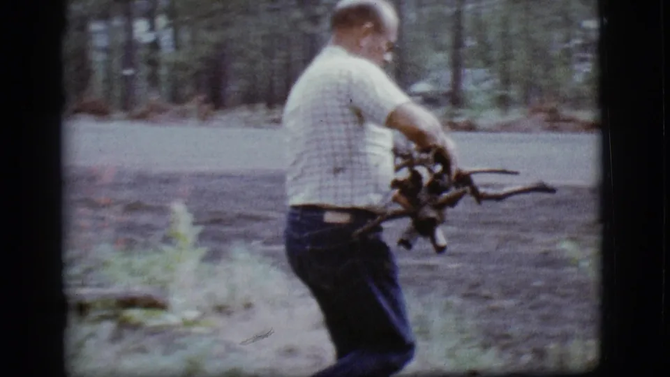 An older man in a light checkered shirt holds gathered sticks near the edge of a paved forest road in 1968 Flagstaff footage.