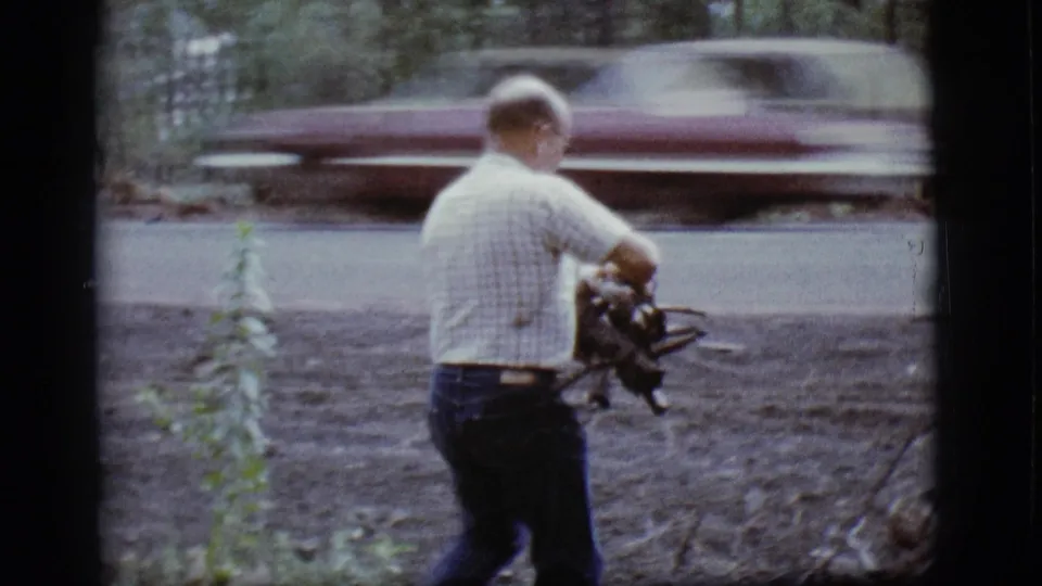 The older man holds a bundle of sticks beside the road while a blurred car passes behind him in the wooded Flagstaff setting.