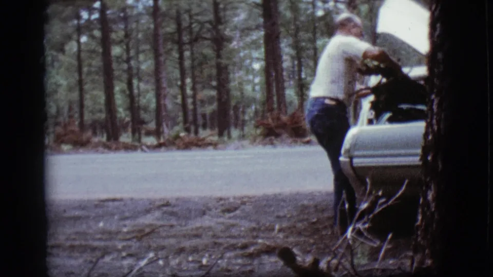 The man reaches the light-colored car with its hood open along a forested roadside in the 1968 home-movie footage.
