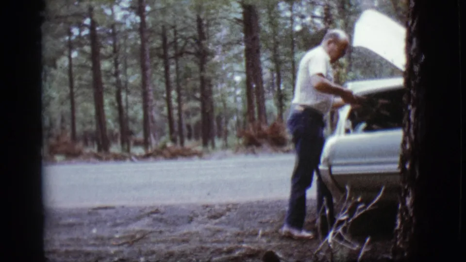 Adult man beside a light-colored car with its hood open on a wooded road in Flagstaff, Arizona, 1968.