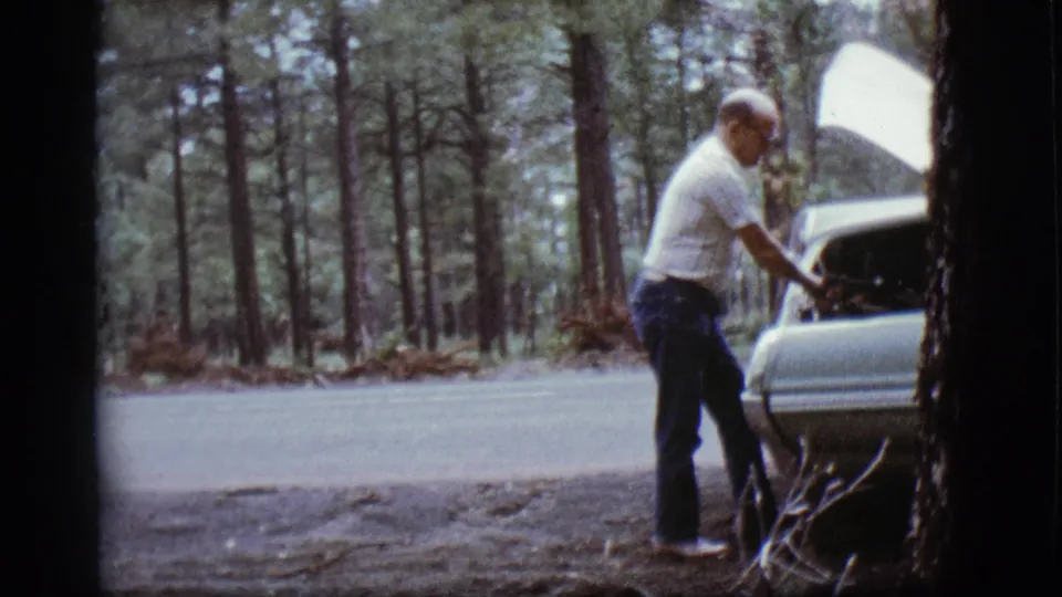 Adult man works near the raised hood of a light-colored car on a wooded Flagstaff roadside in 1968.