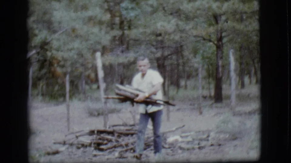 Adult man carries gathered branches through a wooded Flagstaff area in this 1968 8mm home movie frame.