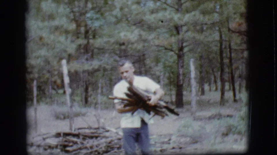 The man carries a bundle of branches through the Flagstaff pine woods, moving closer to the camera in soft 8mm color.