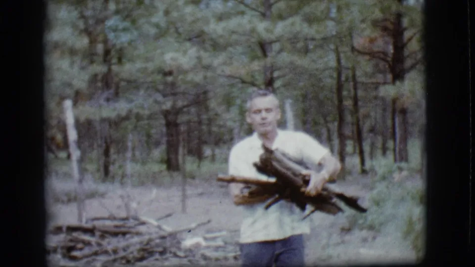 A closer 1968 home-movie frame shows the man carrying firewood toward the camera with a pine forest and woodpile behind him.