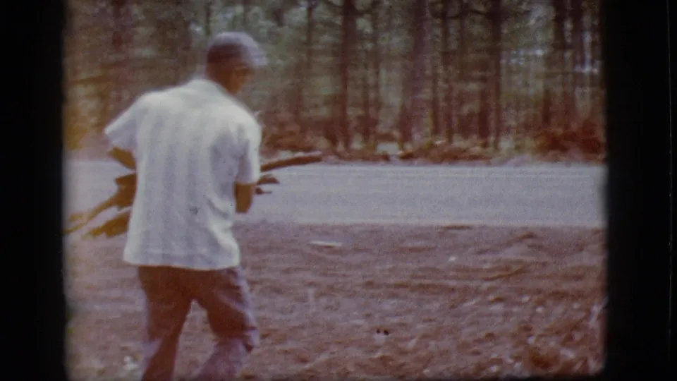 The man walks away carrying firewood across a pine clearing, with warm color fading and dark 8mm frame edges.