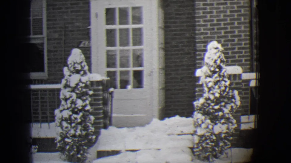 1944 Buffalo 8mm frame of a snow-covered residential porch with a white door, brickwork, railing, and evergreen shrubs.