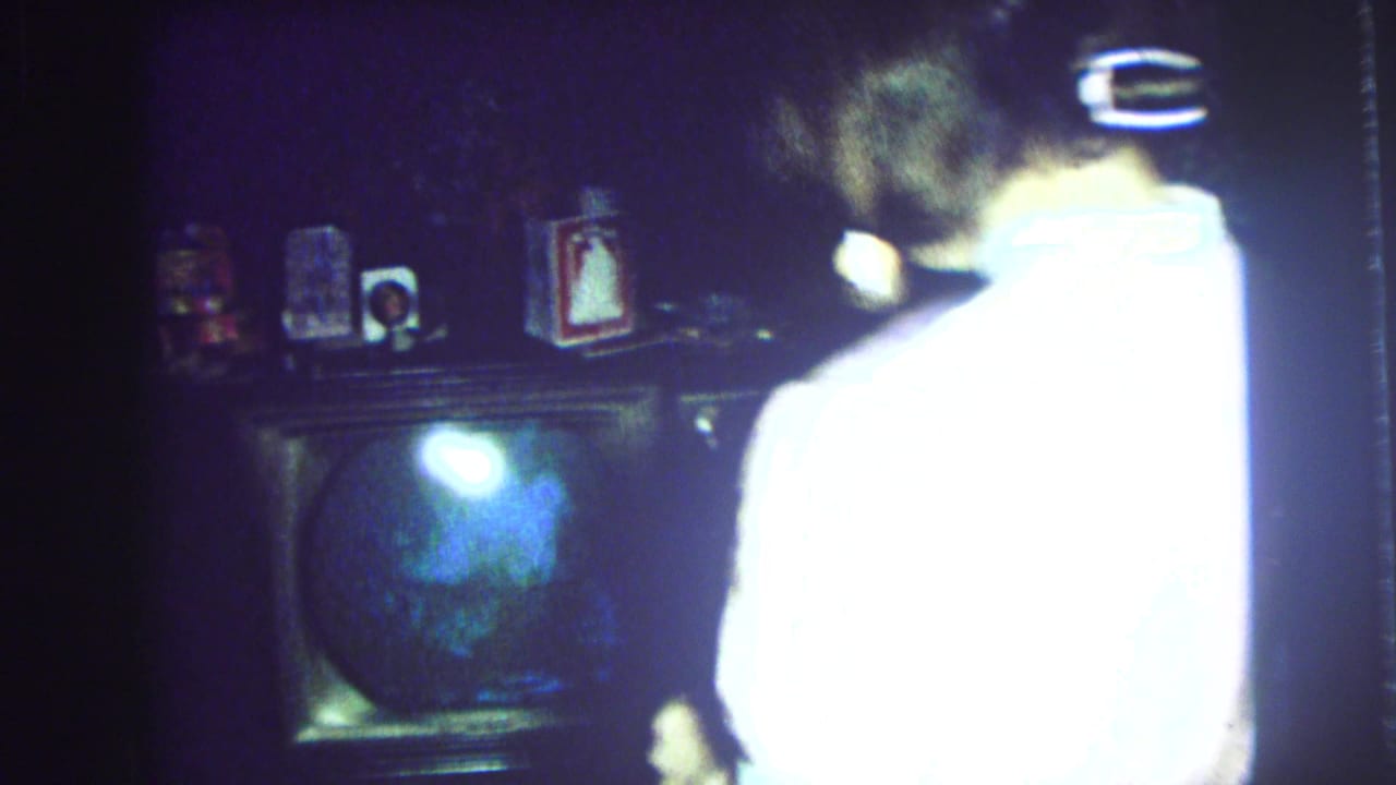 Woman in a light robe stands near a television and mantel in a dark room.