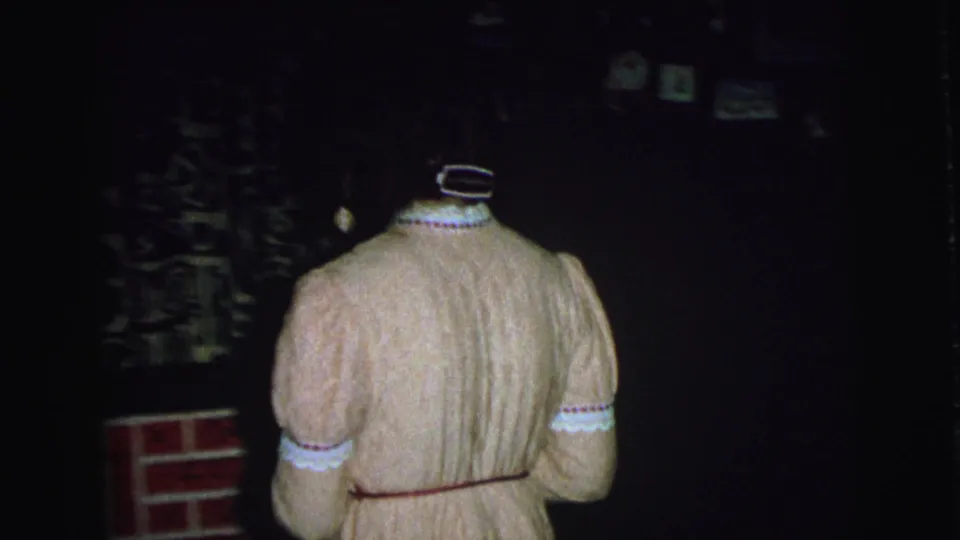 Woman in a pink robe, seen from behind, stands near a fireplace and mantel in a dim 1976 Lynbrook home interior.