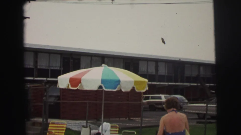 1971 Yellowstone-area poolside scene showing a multicolor umbrella, chain-link fence, lounge chairs, parked car, and a seated person partly in frame.