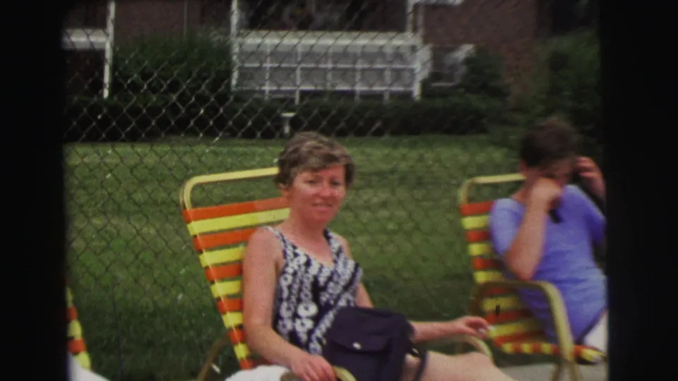 1971 Yellowstone-area home movie frame showing a woman on a yellow-orange striped lounge chair beside another vacationer near a grassy fenced pool area.