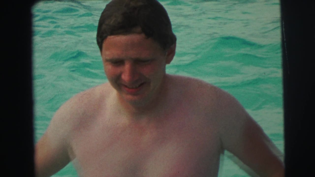 Smiling adult man approaches the camera in a 1971 home-movie pool scene, with turquoise water behind him.