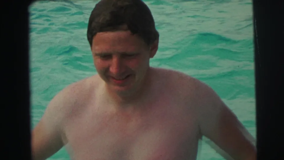 Smiling adult man approaches the camera in a 1971 home-movie pool scene, with turquoise water behind him.