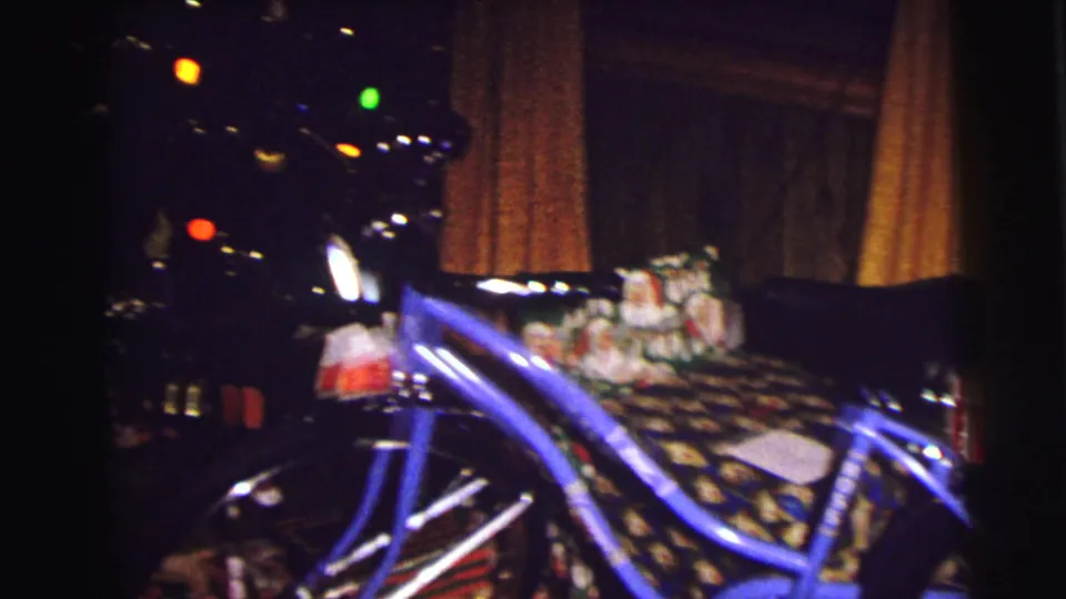 1972 East Lansing home-movie frame showing a blue bicycle, wrapped gifts, and warm Christmas tree lights inside a private room.