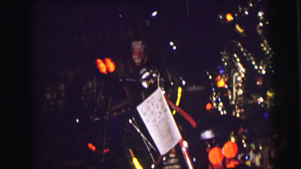 Dark 8mm frame from East Lansing in 1972 with tree lights, tinsel, ornaments, and a hanging paper tag beside bicycle parts.