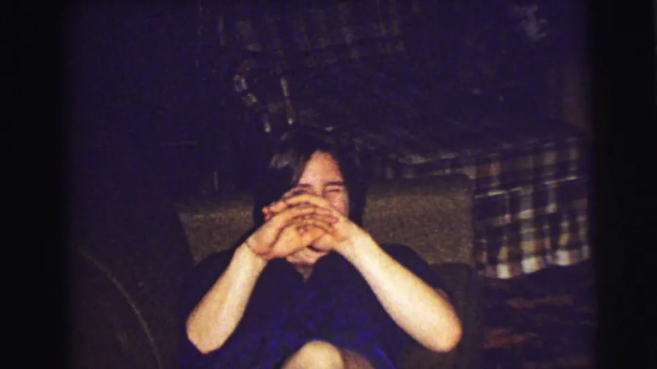A person in a dark shirt covers their face while seated against plaid fabric in a 1972 East Lansing home interior.