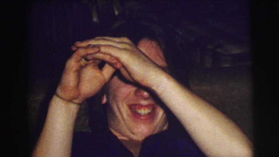 In this East Lansing home-movie frame, the subject covers their eyes while smiling, with a ring visible on one raised hand.