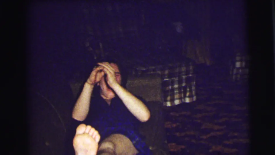 A wider 1972 East Lansing frame shows the seated person hiding their face, with a plaid couch and bare foot visible in the dim room.