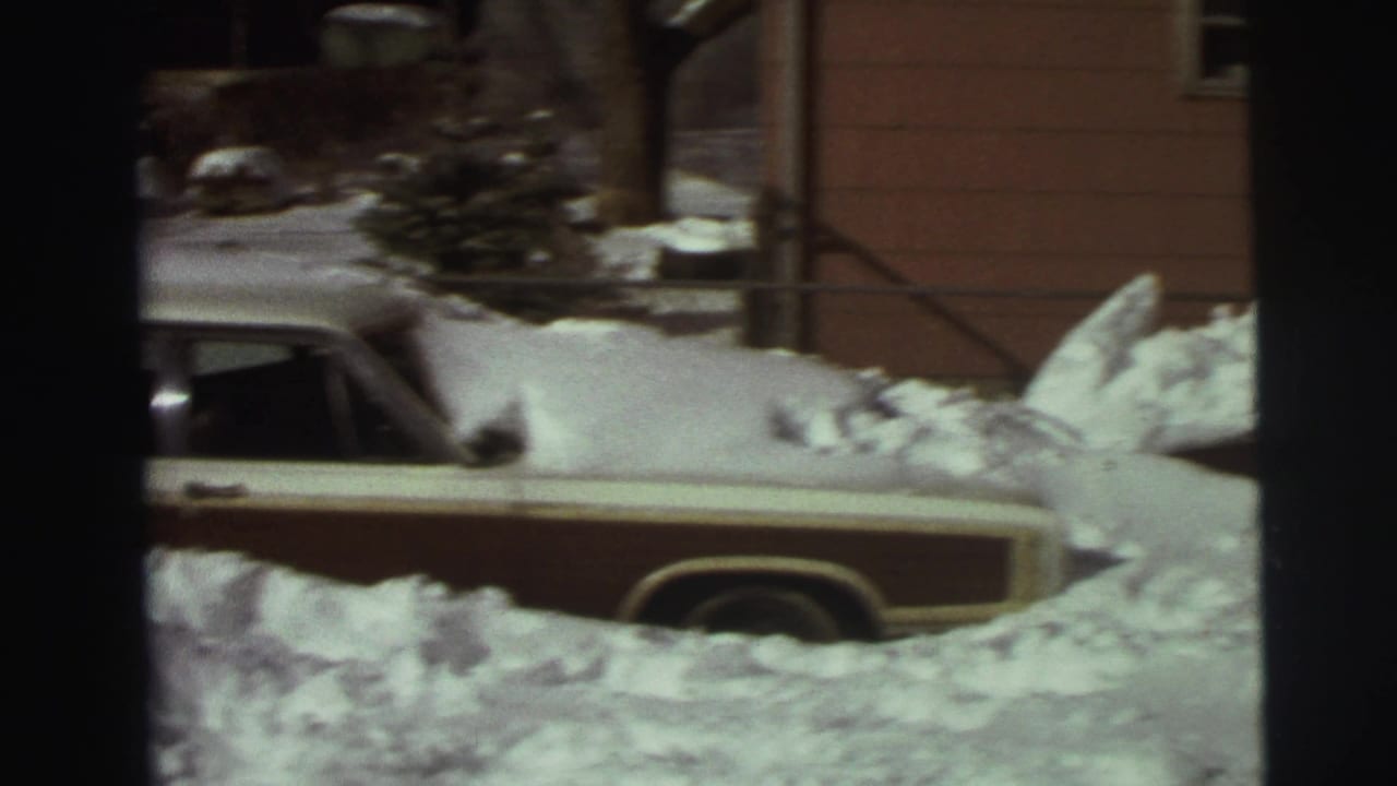 Snowbound driveway with a light-colored station wagon and red sedan near a house.