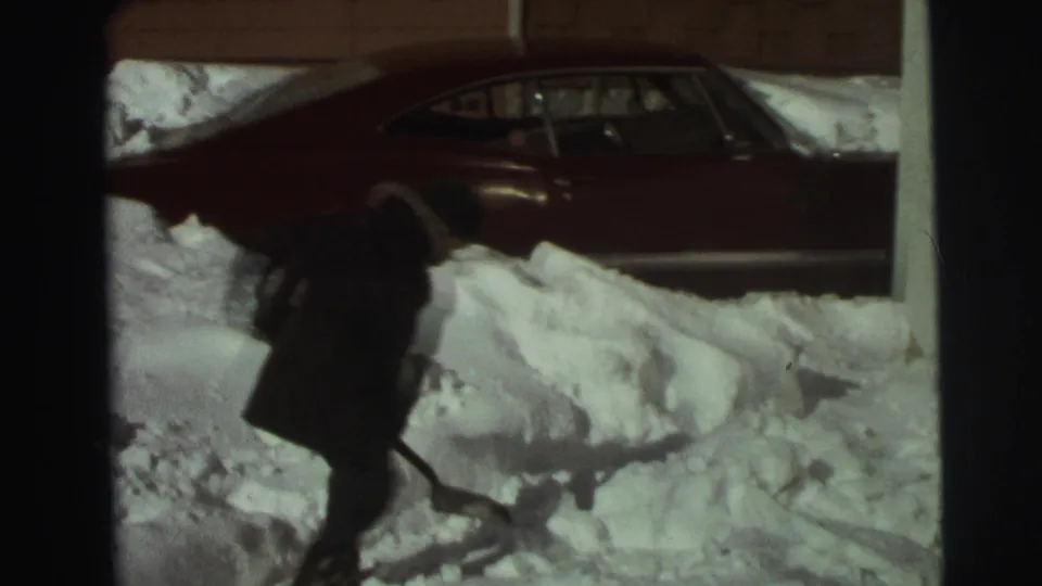 A bundled person shovels deep snow beside a red sedan in 1972 East Lansing home-movie footage.