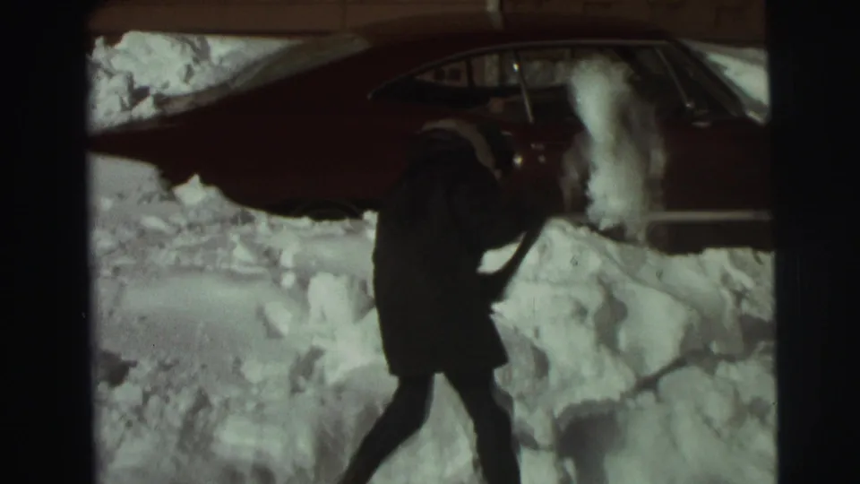 A bundled person lifts or throws snow with a shovel near a buried red sedan in East Lansing, 1972.