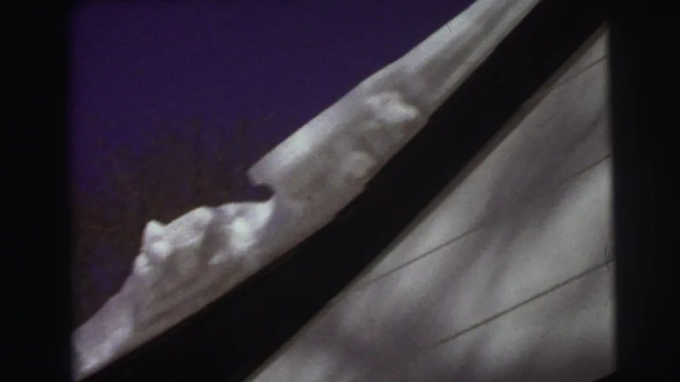 1972 East Lansing frame with thick roof snow, white siding, a dark roofline, and bare winter branches in the background.
