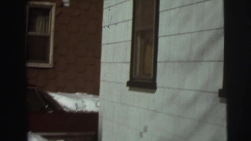 1972 East Lansing residential exterior with white siding, brick wall, windows, and a red car partly covered by snow near the house.