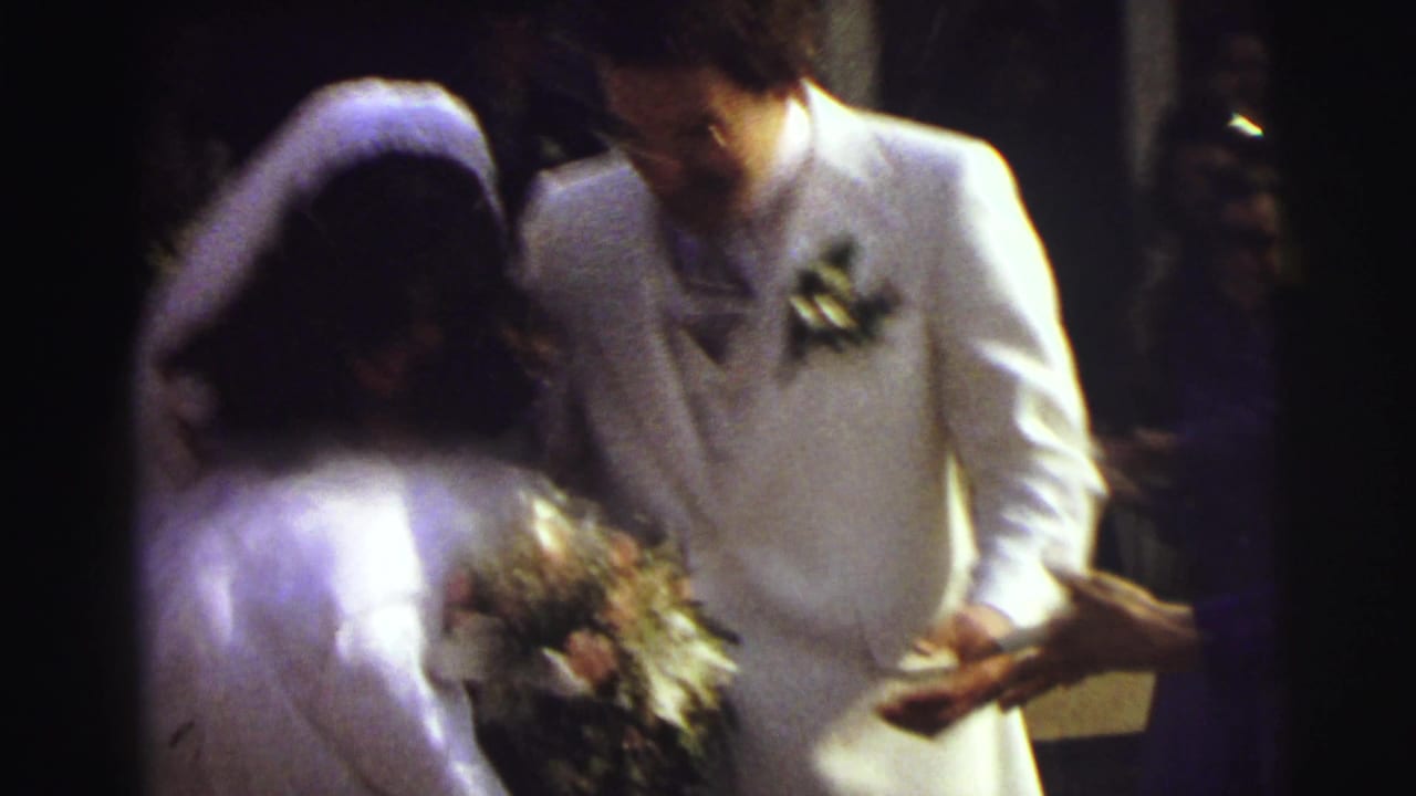 In 1972 East Lansing wedding home-movie footage, a bride in a white veil and a groom in a white suit stand amid guests as rice is thrown around them.