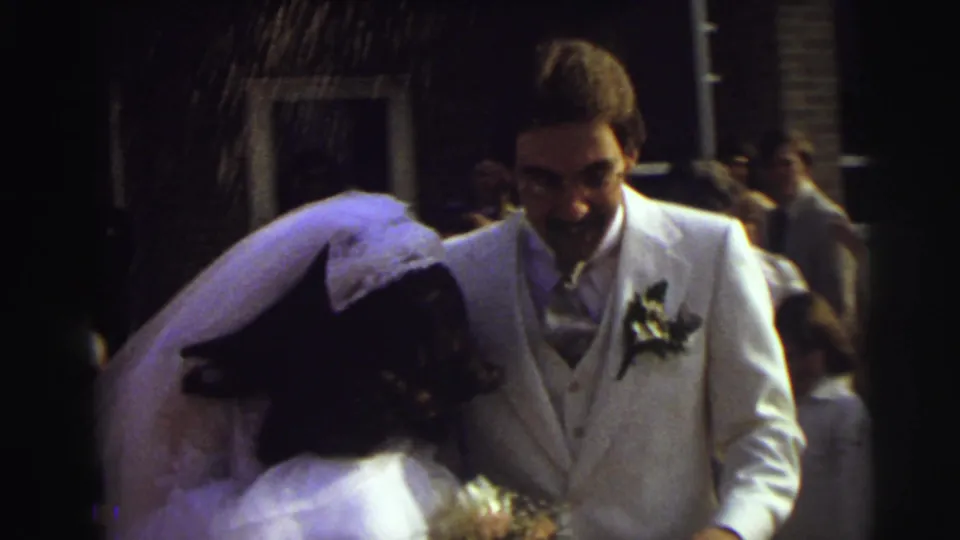 The groom in a white suit smiles toward the veiled bride while guests stand nearby and rice falls through the frame in this 1972 East Lansing wedding scene.