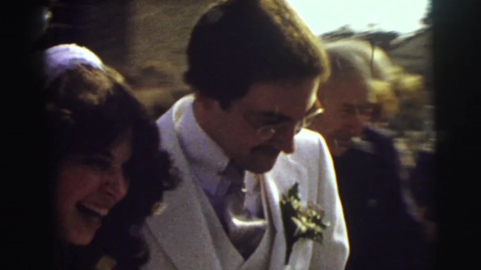 The bride laughs beside the groom in a white suit as guests surround the couple during a handheld 1972 East Lansing wedding exit.