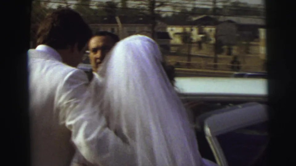 The bride in a long white veil and the groom in a white suit move toward a waiting vehicle as guests watch during the 1972 East Lansing wedding exit.