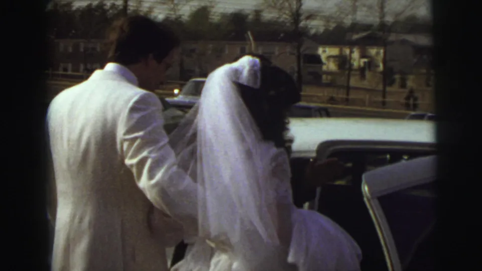 In East Lansing, Michigan in 1972, a bride in a white veil appears to hesitate beside an open car as the groom helps from behind.